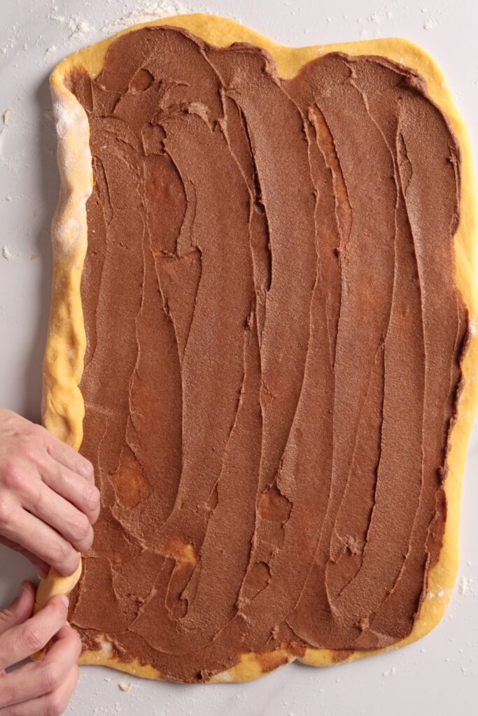 Hands rolling up dough spread with cinnamon filling on a floured surface, preparing to shape it. The dough is rectangular and the filling is smoothly spread, ready to be rolled into a log.