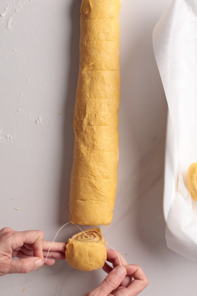 Hands use a piece of string to cut a rolled log of dough into individual pieces on a white surface, with parchment paper and dough rounds nearby.