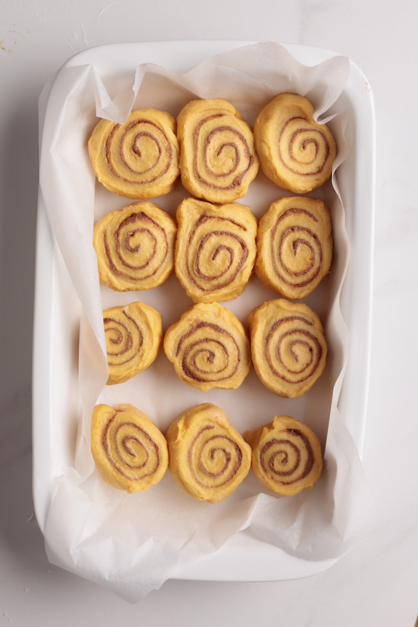Twelve unbaked pumpkin cinnamon rolls arranged neatly in a parchment-lined white baking dish on a light surface.
