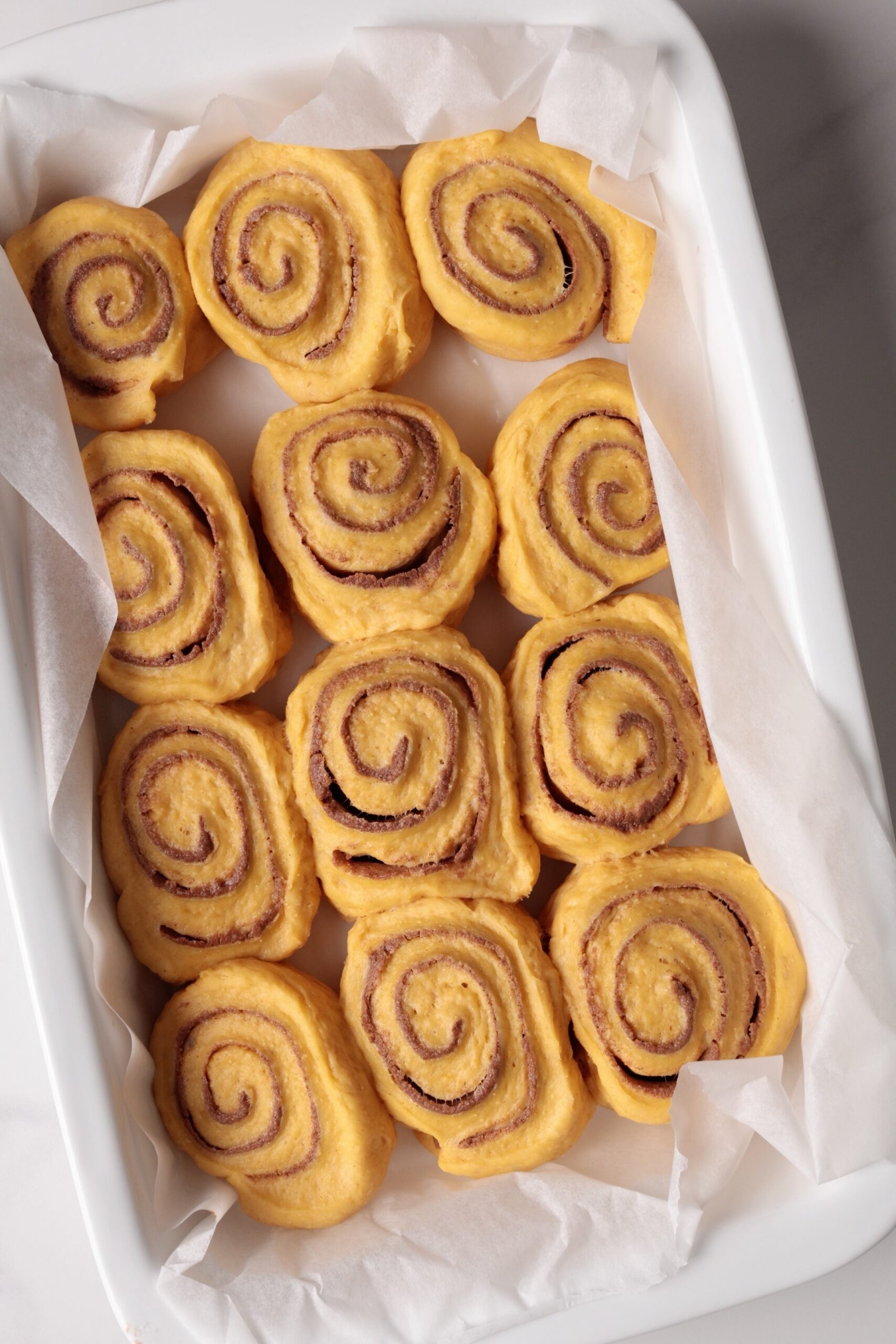 A white baking dish lined with parchment paper, filled with unbaked risen pumpkin spice cinnamon rolls arranged in rows on a light surface.