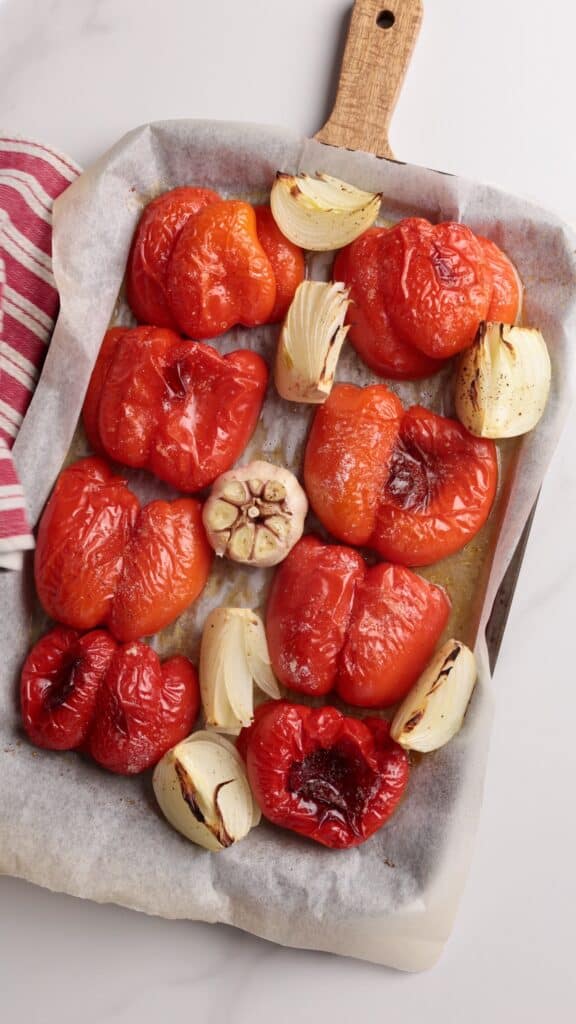 Roasted red bell peppers, onion wedges, and a halved garlic bulb on a parchment-lined baking tray with a wooden handle. A red and white striped cloth is partially visible on the side.