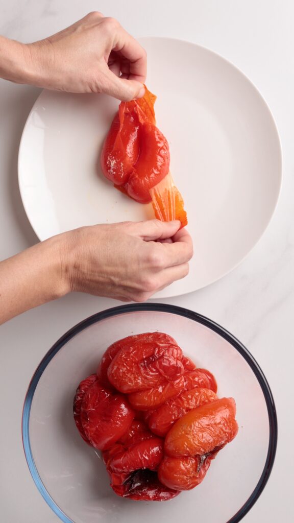 A person peels the skin off a roasted red bell pepper over a white plate, with more peeled peppers in a glass bowl nearby.