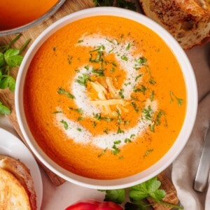 Bowl of creamy roasted pepper gouda soup garnished with cream, shredded cheese, and chopped herbs, surrounded by slices of toasted bread and fresh parsley.