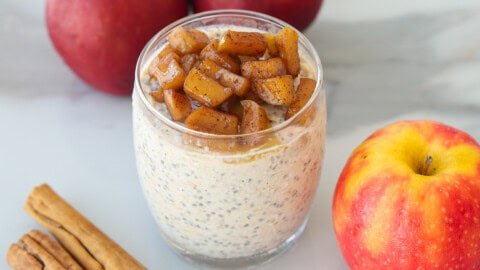 Apple Pie Overnight Oats Glass jar of apple cinnamon overnight oats topped with cooked apple pieces, with whole apples and cinnamon sticks nearby.